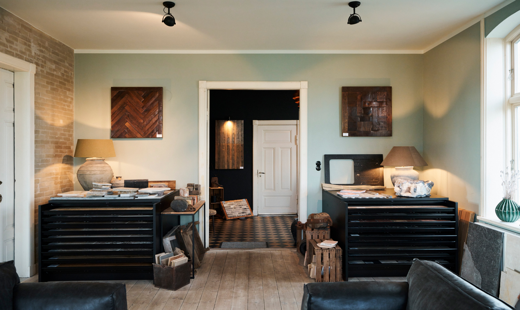 Ansicht von einem großen quadratischen Raum auf die Zimmertür. Rechts und Links von der Tür stehen schwarze Anrichten auf denen jeweils eine beige Lampe und Materialien liegen. Über den Anrichten hängen dunkle Bilder. Durch die Zimmertür kann man in den Flur gucken und sieht eine weitere Tür, diese ist geschlossen.