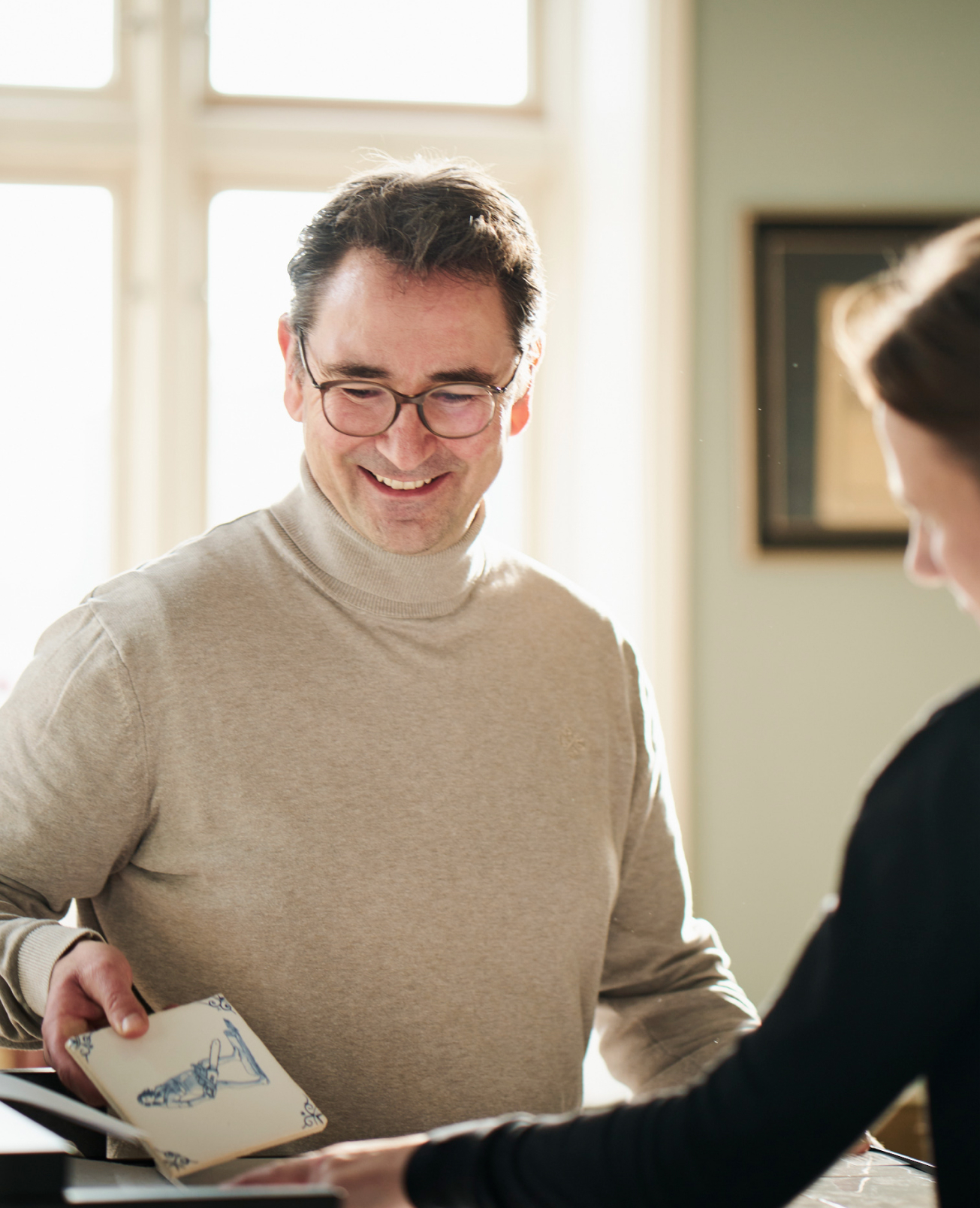 Ein Mann mit einem hellen Pullover, in seiner rechten Hand hält er eine helle Rechteckige Fliese auf der in blaue eine Person zu erkennen ist. Im HIntergrund ist ein Fenster. Im Vordergrund kann man rechts eine Frau erkennen.