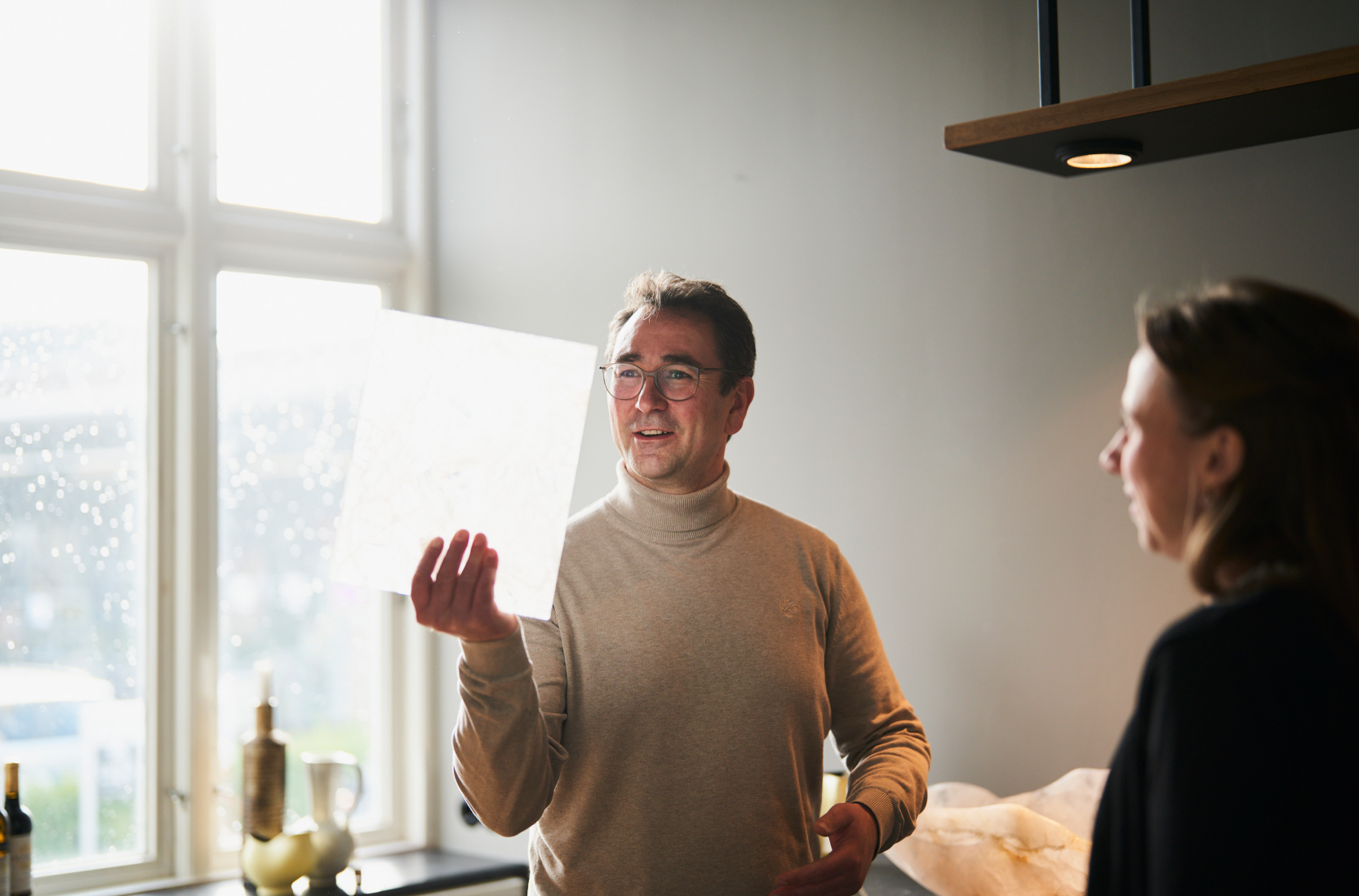 Ein Mann in einem hellen Pullover hält ein Papier ins Licht. Er steht mit dem Rücken zu einem großen Fenster. Im Vordergrund erkennt man eine dunkel gekleidete Frau.